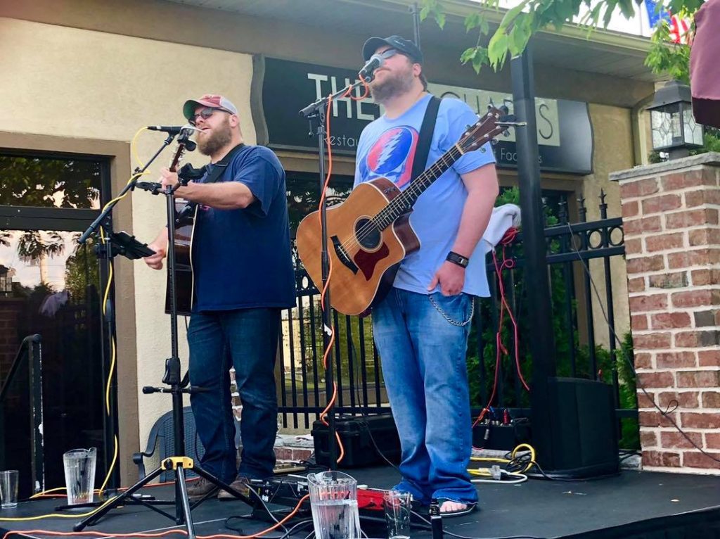 Two musicians play acoustic guitars and sing live on an outdoor stage.