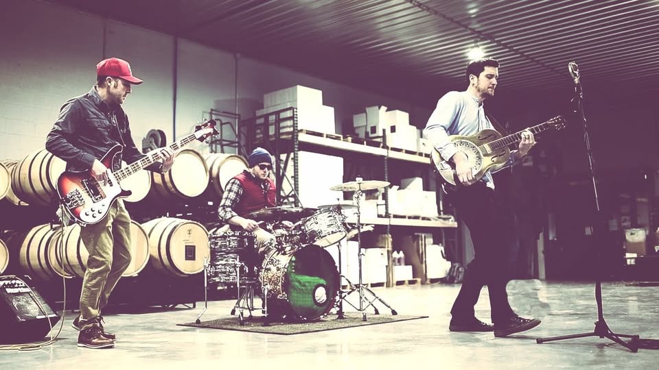 Three musicians perform in a warehouse, with bass, drums, and resonator guitar.