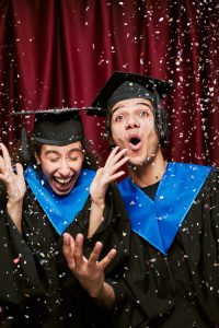 Two young adult graduates, biracial man and multiethnic woman, celebrating in photo booth wearing caps and gowns, smiling and reacting to falling confetti