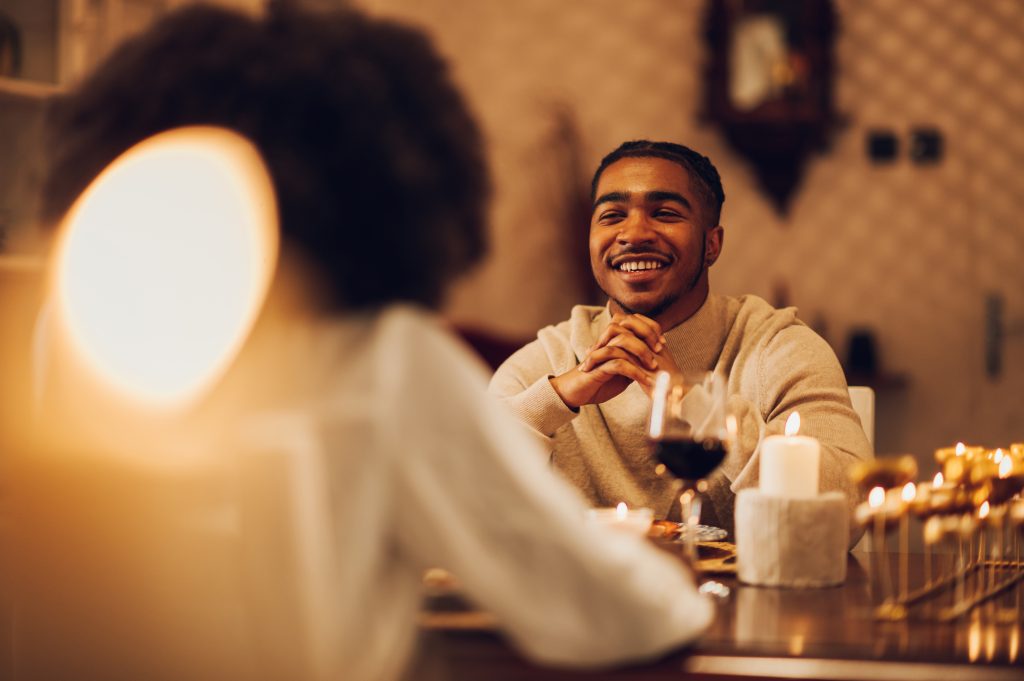 African american married couple having dinner by candlelight at home. Black romantic couple is drinking wine and smiling. Elegant domestic atmosphere. Focus on a diverse man with braids. Copy space.