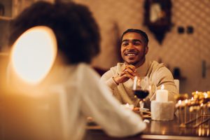 African american married couple having dinner by candlelight at home. Black romantic couple is drinking wine and smiling. Elegant domestic atmosphere. Focus on a diverse man with braids. Copy space.